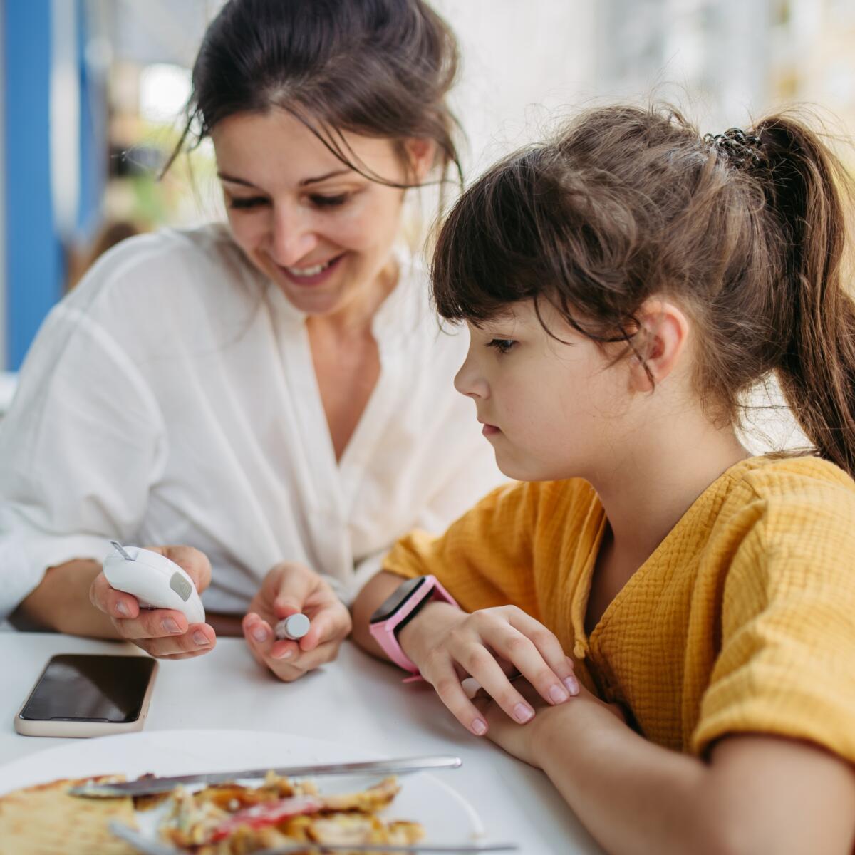 Mother collecting a blood sample from her daughter's finger, using a finger stick test in restaurant. Girl with diabetes checking her blood sugar before eating to plan better her meal.