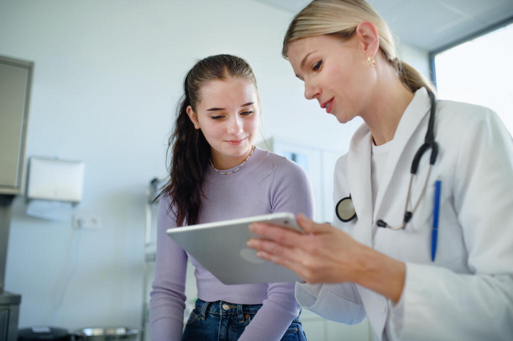 In a doctor’s office, a pediatrician shows a tablet to a teenage patient.